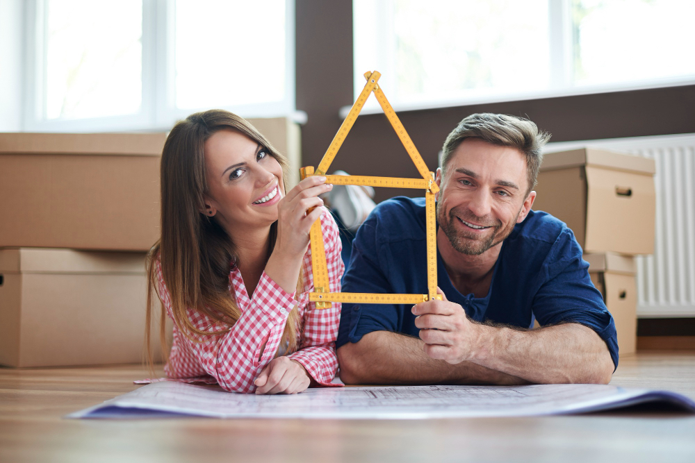 Smiling couple lying on the floor among moving boxes, holding a yellow triangular ruler to outline a house shape over blueprints.