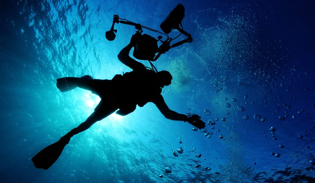 Silhouette of a scuba diver underwater, holding a camera rig as bubbles rise in blue ocean water.