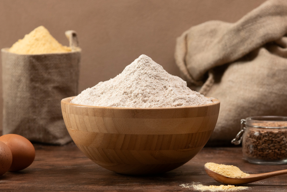 Wooden bowl piled high with white flour on a rustic table, with eggs and burlap sacks in the background for a baking setup.
