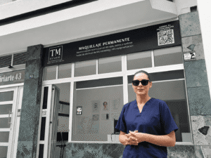 Woman in blue scrubs wearing sunglasses stands outside a beauty clinic offering permanent makeup.