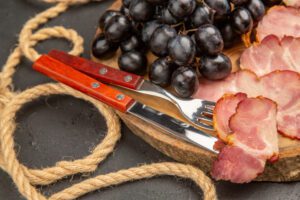 Wooden board topped with sliced cured meat, a bunch of black grapes, and a knife and fork, on a rope-tied surface.