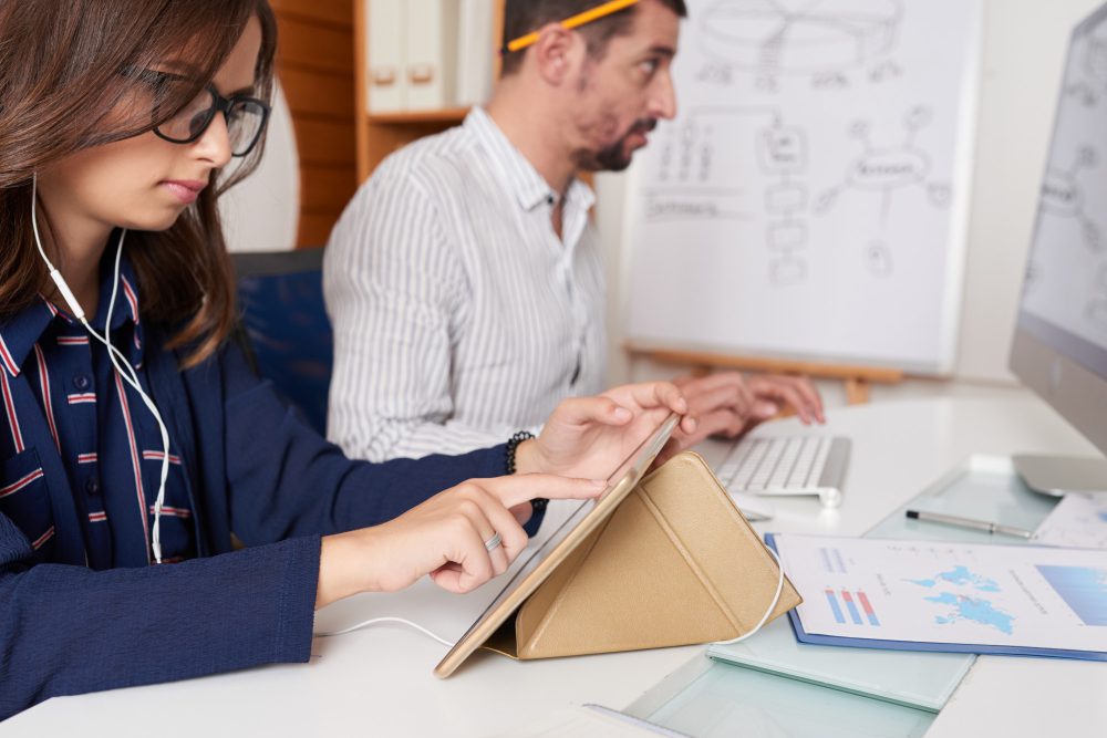 Two coworkers at a bright desk; woman uses a tablet with a case while a man types on a laptop, whiteboard behind them.