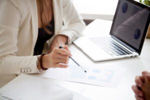 Person in a white blazer analyzes charts on paper with a laptop nearby on the desk.