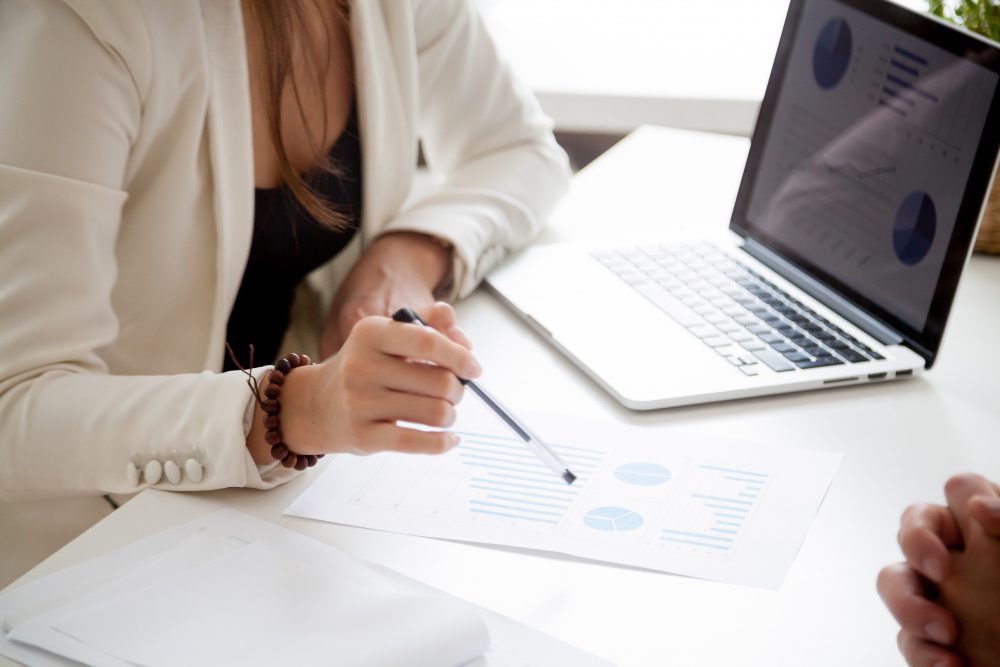 Person in a white blazer analyzes charts on paper with a laptop nearby on the desk.